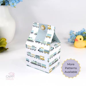 May include: A small white favor box with a blue and green train pattern. The box has a blue ribbon and a gold button on top. The box is perfect for holding small gifts or favors.