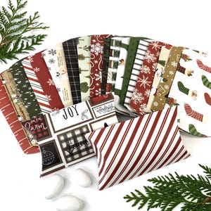 May include: Assortment of Christmas-themed paper gift bags with various patterns, including plaid, snowflakes, and stripes. The bags are folded and arranged in a fan shape. A red and white striped pillow box is in the foreground, with a few white chocolate candies scattered around it.