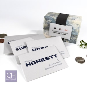 May include: A collection of white cards with navy blue text, including "HONESTY", "HOPE", and "SURI", are arranged in front of a patterned box. The box has a black band and a label that reads "R.P.M. Real Principles for Men Recovery Edition".