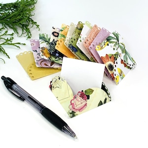 May include: A set of colorful floral patterned paper envelopes with scalloped edges. The envelopes are arranged in a fan shape with a blank white card in the center. A black pen is lying on a white surface.