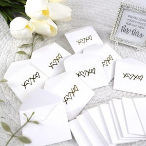 May include: White envelopes with gold foil lettering that says "XOXO" with hearts. The envelopes are on a white fluffy surface. A sign in the background says "Please leave your wishes for the new Mr. & Mrs."
