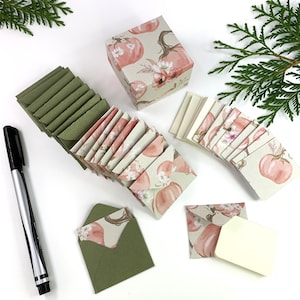 May include: A set of 20 small, square envelopes featuring a floral and pumpkin design. The envelopes are cream coloured with pink and brown accents. The envelopes are packaged in a small, square box with a matching design. The box is also cream coloured with pink and brown accents. A black pen is also included in the image.