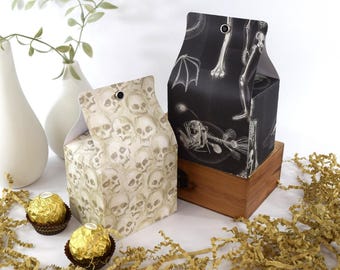 May include: Two decorative gift boxes with a Halloween theme. One box is covered in a skull pattern, and the other features a black background with white illustrations of bones and a fish skeleton. The boxes are on a wooden surface.