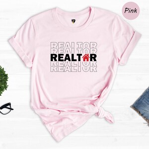 May include: Pink t-shirt with the word "REALTOR" repeated in black text with a red house icon in the middle word. The shirt is on a white wooden background.