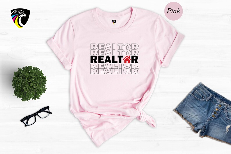 May include: Pink t-shirt with the word "REALTOR" repeated in black text with a red house icon in the middle word. The shirt is on a white wooden background.