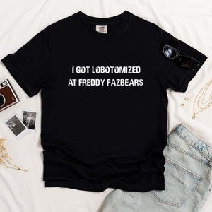 May include: Black t-shirt with the text "I GOT LOBOTOMIZED AT FREDDY FAZBEARS" in white. The shirt is displayed on a white surface with sunglasses, a gold necklace, a vintage camera, and two small photos.