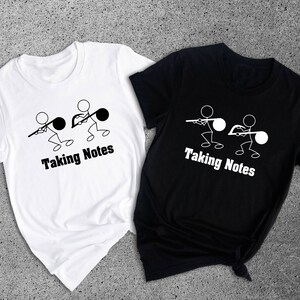 May include: Two white t-shirts with a black stick figure design of two people pulling on a musical note. The text "Taking Notes" is below the design. One t-shirt is white and the other is black.