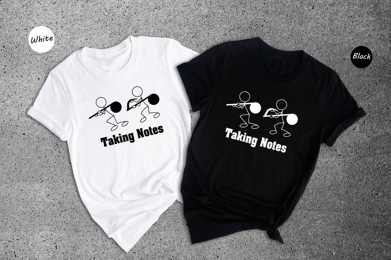 May include: Two white t-shirts with a black stick figure design of two people pulling on a musical note. The text "Taking Notes" is below the design. One t-shirt is white and the other is black.
