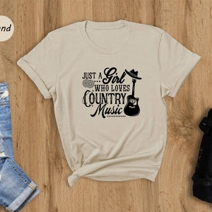 May include: A beige t-shirt with a black graphic that says "Just a Girl... Who Loves Country Music" with a guitar and a cowboy hat.