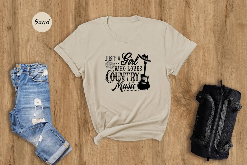 May include: A beige t-shirt with a black graphic that says "Just a Girl... Who Loves Country Music" with a guitar and a cowboy hat.