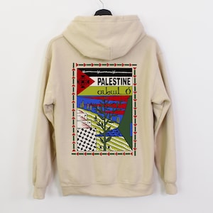 Peut inclure: Un sweat à capuche beige avec un imprimé graphique au dos. Le graphique représente un paysage stylisé avec un arbre, un bâtiment et un drapeau avec le mot "PALESTINE" et du texte en arabe. Le graphique est entouré d'une bordure rouge, verte et bleue.