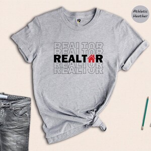 May include: A grey t-shirt with the word "REALTOR" repeated in black text with a red house icon in the centre of the word. The shirt is on a white background.