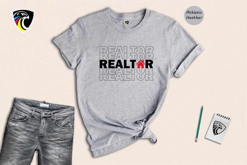 May include: A grey t-shirt with the word "REALTOR" repeated in black text with a red house icon in the centre of the word. The shirt is on a white background.