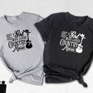 May include: Two t-shirts with a country music design. The gray t-shirt has black text and the dark gray t-shirt has white text. Both shirts feature a guitar and the text "Just a Girl Who Loves Country Music".