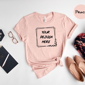 Pode incluir: Camiseta cor p&ecirc;ssego com uma moldura quadrada preta em torno do texto "YOUR DESIGN HERE". A camiseta est&aacute; colocada sobre uma superf&iacute;cie branca com um caderno preto, um rel&oacute;gio, &oacute;culos e um len&ccedil;o floral.