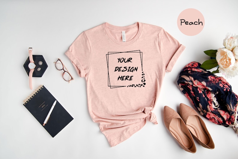 Pode incluir: Camiseta cor p&ecirc;ssego com uma moldura quadrada preta em torno do texto "YOUR DESIGN HERE". A camiseta est&aacute; colocada sobre uma superf&iacute;cie branca com um caderno preto, um rel&oacute;gio, &oacute;culos e um len&ccedil;o floral.