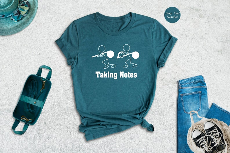 May include: A teal blue t-shirt with a white stick figure design of two people carrying a large note. The text "Taking Notes" is printed below the figures.