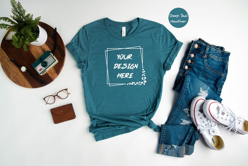 Pode incluir: Uma camiseta de cor azul-esverdeada com o texto "YOUR DESIGN HERE" dentro de um quadro branco. A camiseta est&aacute; colocada sobre uma superf&iacute;cie branca com um par de cal&ccedil;as de ganga, t&eacute;nis brancos e outros acess&oacute;rios.