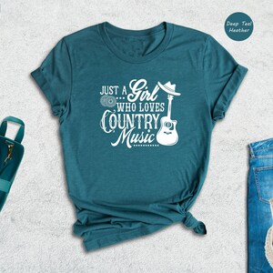 May include: A teal blue t-shirt with the text "Just a... Girl who loves Country Music" in white. A white guitar and cowboy hat are also included in the design.