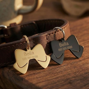 Può includere: Due targhette per animali a forma di fiocco, una dorata e una nera, con il nome "Bella" e un numero di telefono incisi. Le targhette sono attaccate a un collare di cuoio marrone, su una superficie di legno.