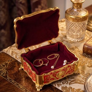May include: A rectangular jewelry box with a gold metal frame and red decorative panels, featuring a hinged lid and a deep red velvet interior lining. Inside the box are two gold rings, a delicate gold chain necklace, and two pearl stud earrings.