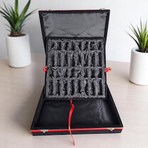 Può includere: Scatola da scacchi nera aperta con finiture rosse e un nastro rosso. L'interno è foderato in tessuto nero e ha un inserto sagomato per contenere i pezzi degli scacchi. Due piccole piante in vasi bianchi sono sullo sfondo.