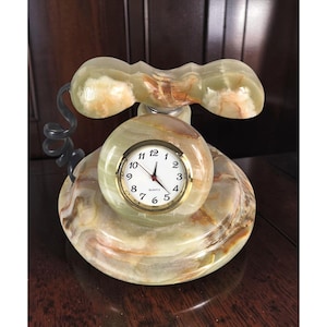 May include: A decorative desk clock crafted from polished green and brown banded onyx, shaped like a vintage telephone. The clock face is circular with a gold-tone rim, black numerals, and three hands, featuring the word Quartz printed on the dial.