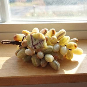 May include: A decorative cluster of grapes made from polished stone. The grapes vary in color from pale yellow to green and white, with a brown stem. The grapes are arranged on a wooden surface near a window.