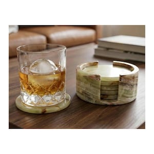 May include: A clear glass tumbler with a diamond-cut pattern containing an amber liquid and a large spherical ice cube, resting on a round green onyx coaster next to a matching cylindrical stone coaster holder containing additional coasters.
