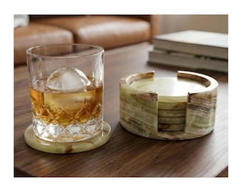 May include: A clear glass tumbler with a diamond-cut pattern containing an amber liquid and a large spherical ice cube, resting on a round green onyx coaster next to a matching cylindrical stone coaster holder containing additional coasters.
