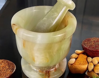 May include: A green and brown marble mortar and pestle set. The mortar is a bowl shape with a wide base and the pestle is a smooth, rounded cylinder. The set is sitting on a black surface with almonds and spices around it.