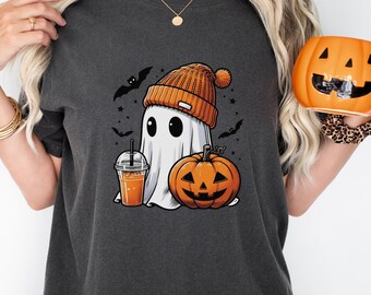 Puede incluir: Una camiseta gris carbón con un gráfico de un fantasma blanco que lleva un gorro de punto naranja, sostiene una bebida helada naranja en un vaso de plástico transparente y está junto a una calabaza tallada naranja con murciélagos negros.