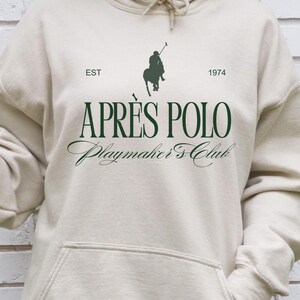 Puede incluir: Sudadera con capucha beige con cordón y bolsillo de canguro. En la parte delantera, el texto "APRÈS POLO Playmaker's Club" en verde oscuro, un logotipo de jugador de polo y el año "1974". La sudadera está hecha de un material suave y cómodo.