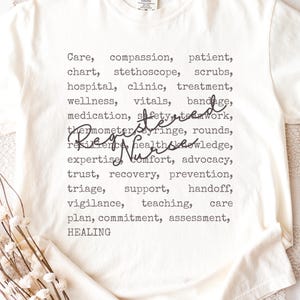 May include: Cream-colored t-shirt with a list of words related to nursing, including "Registered Nurse" in cursive script. The words are printed in a dark gray font. The shirt is made of a soft material.