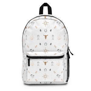 May include: A white backpack featuring a repeating pattern of tan suns, brown bull skulls, black horseshoes, black cacti, and tan arrows. The bag includes a front zippered pocket, a black top handle, and a black mesh side pocket for storage.