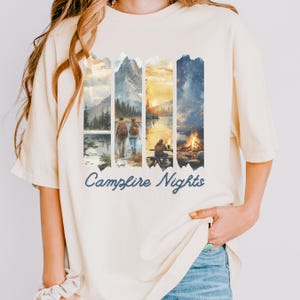 Könnte beinhalten: Cremefarbenes T-Shirt mit einem Aquarelldesign von Berglandschaften und einer Lagerfeuerszene. Das Shirt trägt den Schriftzug "Campfire Nights" in Schreibschrift. Das Design zeigt Bilder von Wanderern und einem Lagerfeuer.