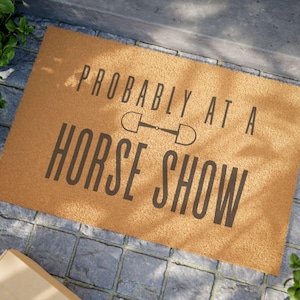 May include: A rectangular doormat with the text "PROBABLY AT A HORSE SHOW" in dark gray. The mat is a light brown color and features a horse bit graphic between the words. The mat is placed on a gray stone surface.