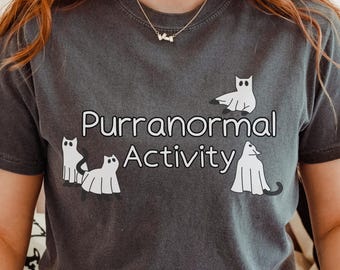 May include: Dark gray t-shirt with the phrase "Purranormal Activity" in white, accompanied by cartoon ghost cats. The shirt is a casual style, suitable for everyday wear. The design is playful and themed around Halloween.