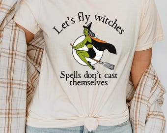 Puede incluir: Camiseta color crema con una bruja de dibujos animados montada en una escoba dentro de un círculo blanco. La bruja es verde con un sombrero y capa negros. El texto dice "Let's fly witches" y "Spells don't cast themselves."
