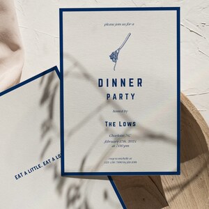 Op de afbeelding: Een witte uitnodigingskaart met een donkerblauwe rand. De kaart is voor een diner dat wordt georganiseerd door The Lows in Charlotte, North Carolina, op 17 februari 2024 om 19:00 uur. De tekst "EAT A LITTLE. EAT A LOT." staat gedrukt op de achterkant van de kaart.