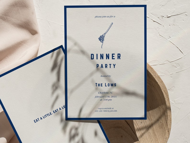 Op de afbeelding: Een witte uitnodigingskaart met een donkerblauwe rand. De kaart is voor een diner dat wordt georganiseerd door The Lows in Charlotte, North Carolina, op 17 februari 2024 om 19:00 uur. De tekst "EAT A LITTLE. EAT A LOT." staat gedrukt op de achterkant van de kaart.