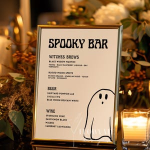 Könnte beinhalten: Ein goldgerahmtes Schild mit der Aufschrift "SPOOKY BAR", das Halloween-Drinks und eine Cartoon-Geisterillustration auflistet. Das Schild ist von Kerzen und Blumengestecken umgeben.