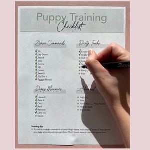 May include: A handwritten puppy training checklist with sections for basic commands, party tricks, puppy manners, and advanced training. A hand holding a black Sharpie pen is checking off items on the list. The checklist is on a white paper with a pink background.