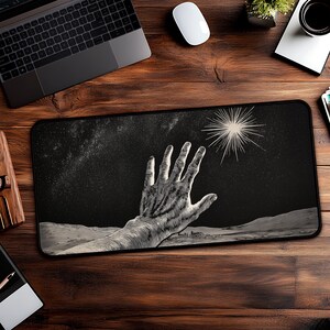 May include: Black rectangular mouse pad featuring a detailed black and white illustration of a hand reaching towards a bright star in a starry night sky. The design includes a landscape at the bottom. The mouse pad is on a wooden desk.