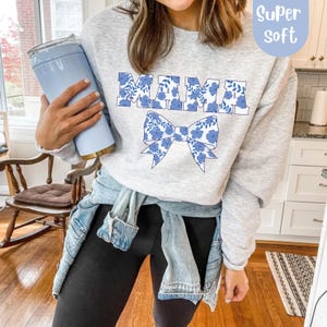 Könnte beinhalten: Ein hellgraues Sweatshirt mit dem Wort "MAMA" in blau floral bedruckten Buchstaben und einer passenden Schleifengrafik. Das Sweatshirt hat einen Rundhalsausschnitt und lange Ärmel. Die Worte "Super Soft" stehen in einem blauen Kreis oben rechts.