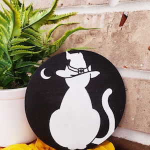May include: A black and white wooden circle with a white silhouette of a cat wearing a witch's hat. The cat is facing away from the viewer and has a crescent moon behind it.