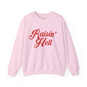 以下が含まれることがあります： 前面に赤い筆記体で「Raisin' Hell」と書かれた、薄いピンク色のスウェットシャツ。クルーネックと長袖のデザインです。テキストは筆記体フォントです。