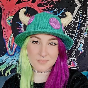 May include: A person with green and purple hair is wearing a green crocheted hat with horns and purple polka dots. The hat is a recreation of the character Mike from the movie Monsters Inc.