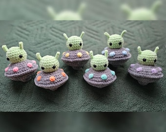 Könnte beinhalten: Sieben gehäkelte grüne Alien-Figuren mit großen Augen und Antennen, die jeweils auf einem kleinen gehäkelten Raumschiff mit bunten Punkten sitzen.