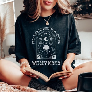 May include: A black t-shirt with a white graphic design featuring a crescent moon, stars, mushrooms, and the text "Pray with me don't play with me Witchy Mama".